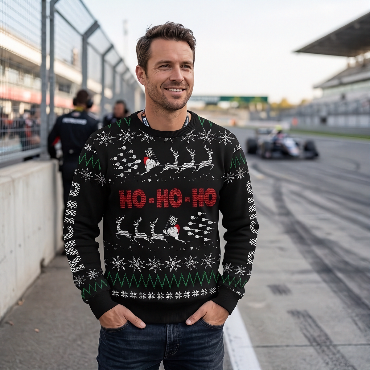 A man wearing a black Christmas sweater with a festive holiday pattern stands on a racetrack. The sweater features 'HO-HO-HO' in red text, white reindeer silhouettes, and green snowflakes.
