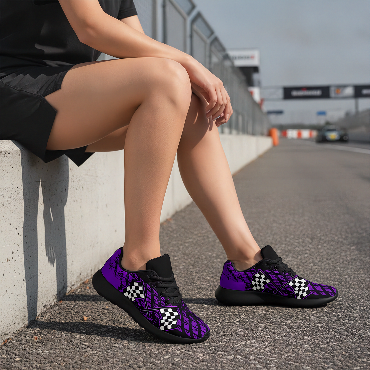 A person sitting on a concrete barrier at a racetrack wearing purple and black patterned sneakers with white checkered accents.