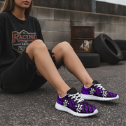 A person sitting outdoors wearing purple and black snakeskin-patterned sneakers with white laces and white soles, paired with a black graphic t-shirt and black shorts.