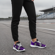 A person standing on an overcast racetrack wearing black jogger pants and purple athletic sneakers with a black and white checkerboard pattern and white laces.