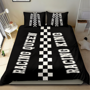 A black bedding set on a bed in a bedroom, featuring a duvet cover and two pillowcases with white racing-themed graphics. The duvet cover displays 'RACING QUEEN' and 'RACING KING' in large white letters separated by a central black-and-white checkered stripe.