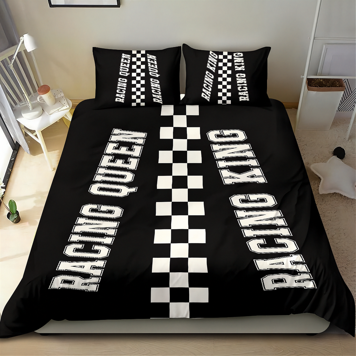 A black bedding set on a bed in a bedroom, featuring a duvet cover and two pillowcases with white racing-themed graphics. The duvet cover displays 'RACING QUEEN' and 'RACING KING' in large white letters separated by a central black-and-white checkered stripe.