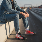 Person wearing red and white checkered shoes sitting on a concrete barrier by a road.