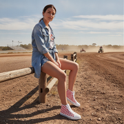 Woman wearing checkered high top shoes sitting on a wooden post with a dirt track and people on motorcycles in the background