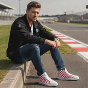Man sitting on a race track wearing a black jacket, blue jeans, and red checkered shoes.