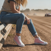 Person wearing red and white checkered shoes sitting on a dirt track with cars in the background.