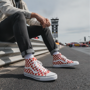 Person wearing red and white checkered high-top shoes sitting on a curb with a race track in the background.