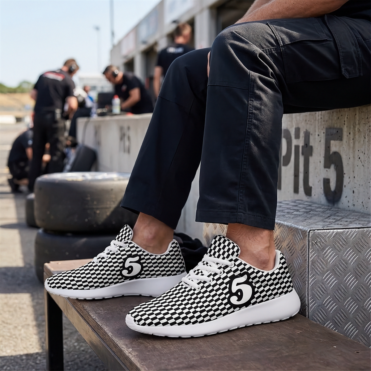 A person sitting on a concrete pit wall at a racetrack wearing black and white checkered sneakers with a large number 5 logo on the side. The background shows race track elements like tires and other people in the pit area.