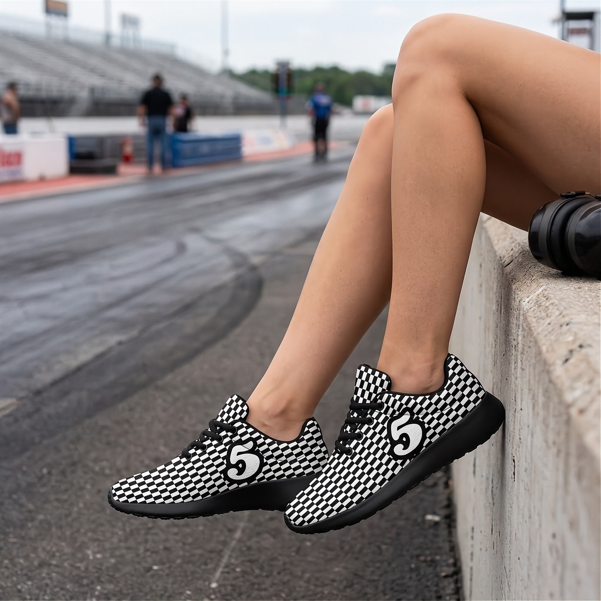 A person sitting on a concrete barrier at a racetrack wearing black and white patterned sneakers with a large white number 5 on the side.