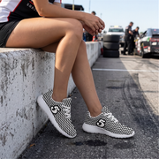A person sitting on a concrete barrier at a racetrack wearing black and white patterned sneakers with white soles, photographed outdoors with cars and people blurred in the background.