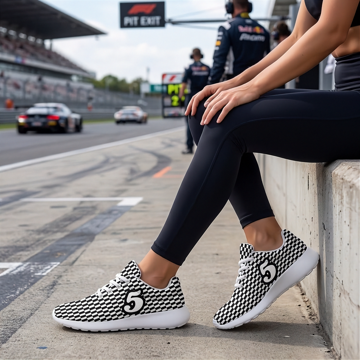 A person sitting on a concrete barrier at a racetrack wearing black and white patterned sneakers with white soles. The background shows a blurred race track, pit crew, and grandstands.