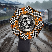 A person holding a large racing-themed umbrella in a muddy, rainy racetrack setting. The umbrella features a black and white checkered pattern, orange accents, and a central silver ring with the word 'Racing' in white script.
