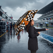 A woman standing on a wet racetrack in the rain, holding an open umbrella with a black-and-white checkered pattern and orange racing graphics. Race grandstands, track barriers, and other umbrellas are visible in the background.