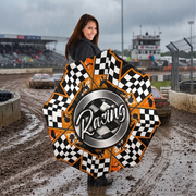 A woman stands on a muddy race track holding a large, star-shaped umbrella with a racing theme. The umbrella features black and white checkered patterns, orange accents, and a central silver circle with the word 'Racing'.