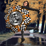 A woman outdoors on a wet road in autumn, holding a large open umbrella with a racing theme. The umbrella features a black and white checkered pattern, orange accents, and a central silver circle with the word 'Racing' in white script.