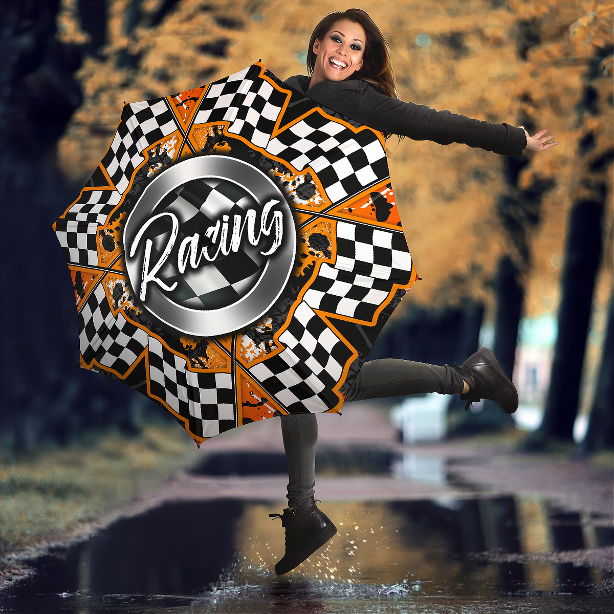 A woman outdoors on a wet road in autumn, holding a large open umbrella with a racing theme. The umbrella features a black and white checkered pattern, orange accents, and a central silver circle with the word 'Racing' in white script.
