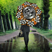 A person walking in the rain on a tree-lined path holding a large racing-themed umbrella. The umbrella features a black-and-white checkered border, orange accents, and a central metallic silver circle with the word 'Racing' in script.