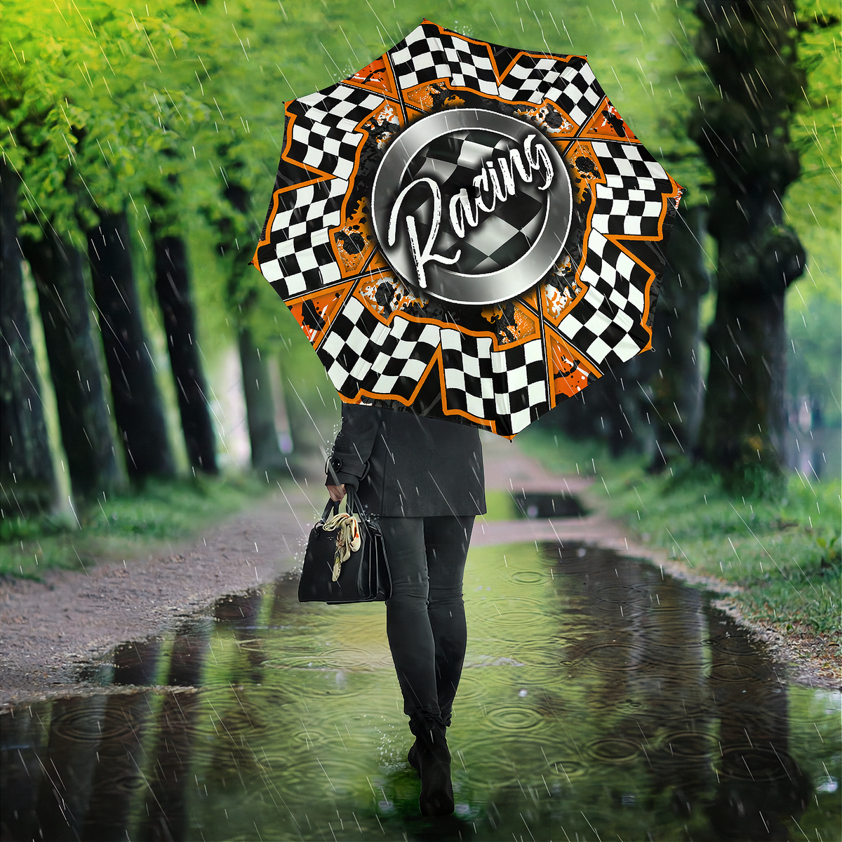 A person walking in the rain on a tree-lined path holding a large racing-themed umbrella. The umbrella features a black-and-white checkered border, orange accents, and a central metallic silver circle with the word 'Racing' in script.