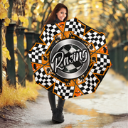 A woman standing on a wet path in an autumn park holds a large, open umbrella. The umbrella features a racing theme with a central silver circle containing the word 'Racing' in white script, surrounded by segments of black and white checkered flags and bright orange sections with small black racing silhouettes.