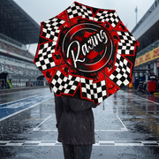A woman in a dark gray coat stands in the rain at a racetrack holding an open umbrella with a black and white checkered pattern and red accents. The racetrack and grandstands are visible in the background under a cloudy sky.