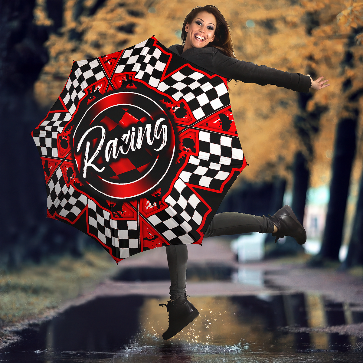 A woman jumps in a puddle outdoors holding a large racing-themed umbrella. The umbrella features a black and red design with checkered segments, a central circular logo reading 'Racing', and small skull graphics.