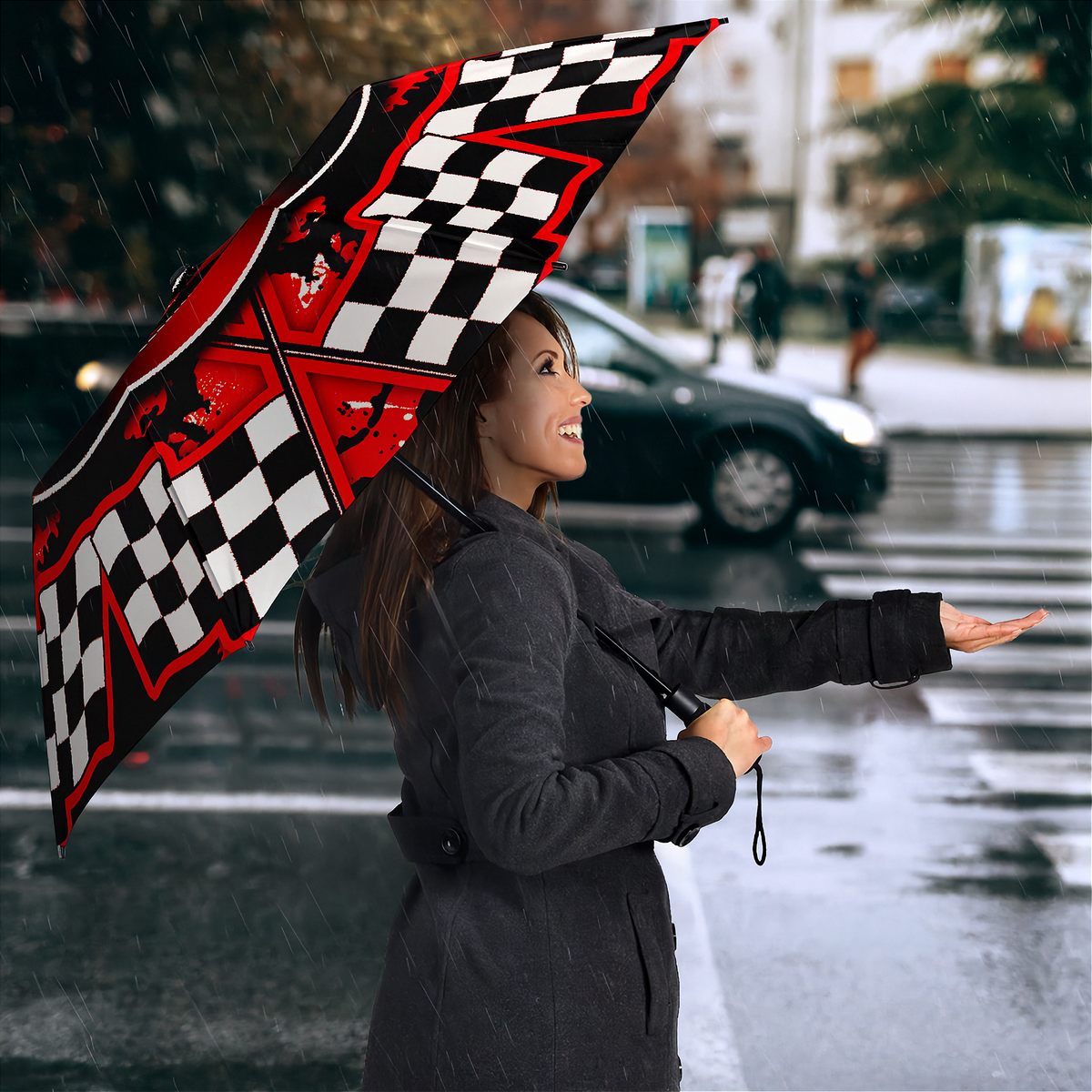A woman in a dark gray coat stands in the rain holding a large open umbrella with a black, white, and red checkered flag design and racing car graphics.