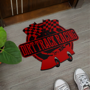 A red and black 'Dirt Track Racing' graphic mat placed on a gray wood-look floor, with a green plant on the left and a person's white sneakers visible at the bottom right.