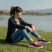 Woman sitting on grass by a lake wearing red and black checkered sneakers.