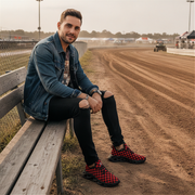 Man sitting on a wooden bench by a race track wearing checkered shoes.