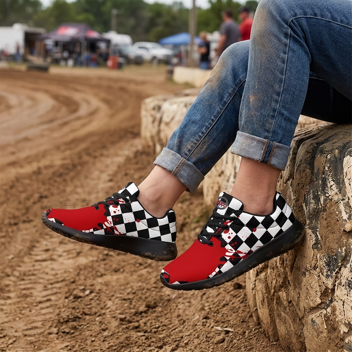 A person sitting on a stone wall at an outdoor dirt track wearing red sneakers with a black and white checkered pattern and cartoon character graphics, paired with cuffed blue jeans.