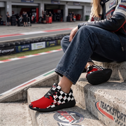 A woman sitting on stairs at a race track wearing red sneakers with a black and white checkered pattern, paired with cuffed blue jeans