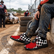 A person sitting at a dirt track wearing red and black checkered racing shoes with distressed detailing, next to stacked tires and a race car in the background.