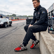 A man in a black racing suit sits on a concrete barrier at a racetrack, wearing red and black checkered high-top sneakers. A classic race car is visible in the background on the track.