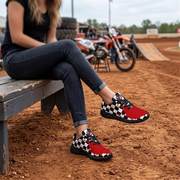 A person sitting on a wooden bench at a dirt track wearing red sneakers with a black and white checkered pattern. An orange dirt bike is blurred in the background.