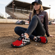 A woman sits on a dirt racetrack wearing a black racing cap, a dark brown racing jacket, and red sneakers with a black-and-white checkered pattern on the sides. Race cars and grandstands are visible in the background.