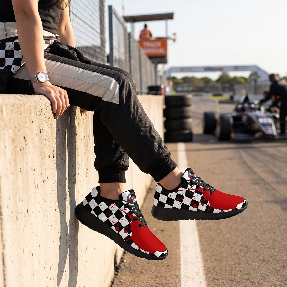 A person sitting on a concrete barrier at a racetrack wearing red, black, and white checkered sneakers, black track pants, and a watch.