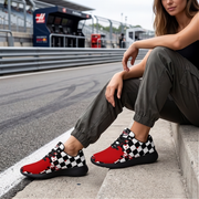 A woman sitting on stairs at a race track wearing red sneakers with a black and white checkered pattern, paired with cuffed grey jeans
