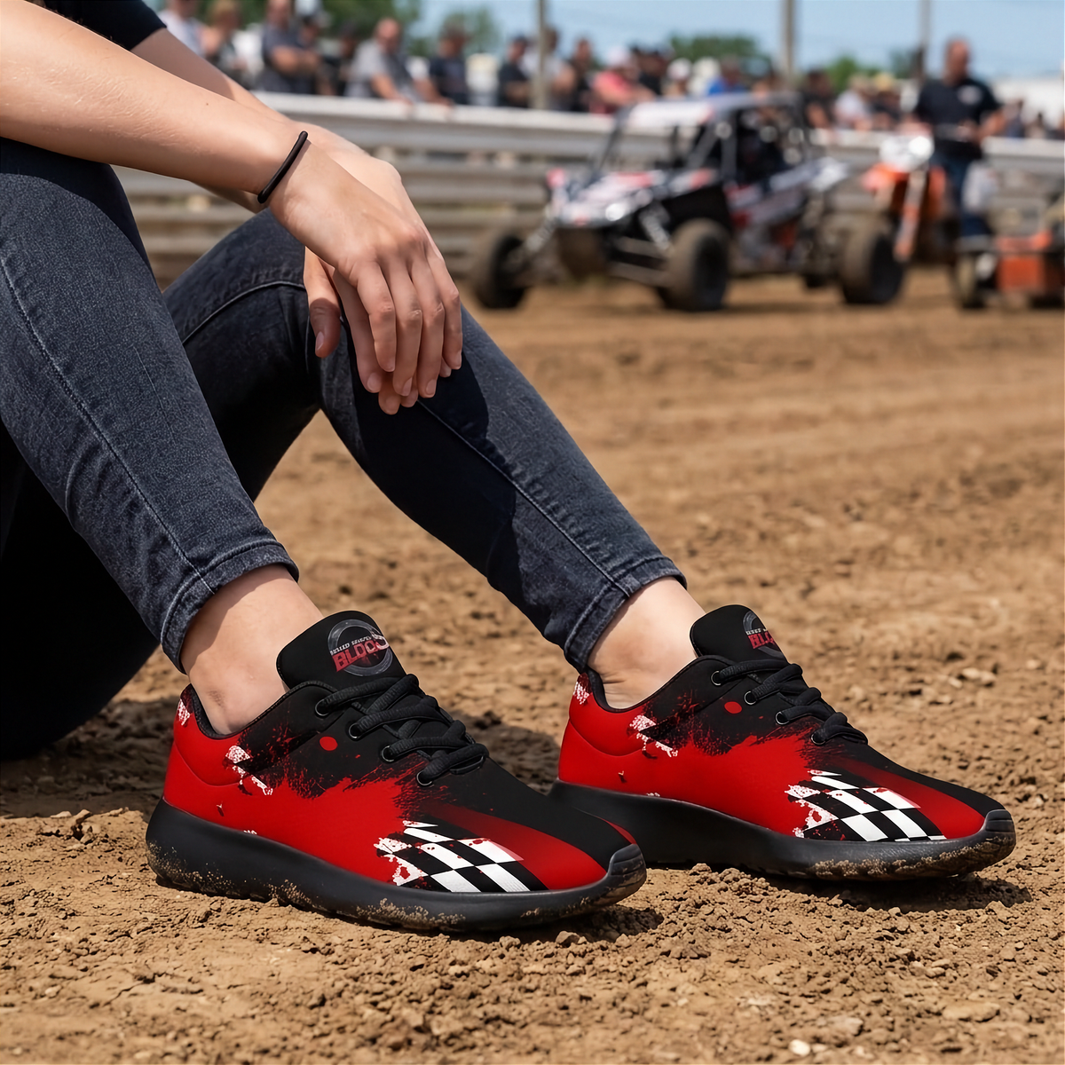 A person sitting on a dirt racetrack wearing red and black checkered sneakers with black jeans, with race cars blurred in the background.