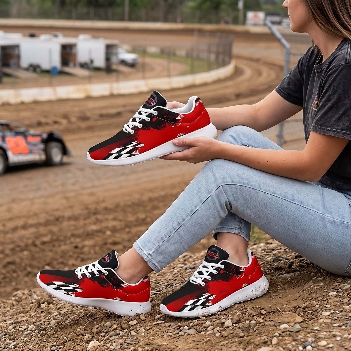 A woman sitting on a dirt track holds up a red, black, and white sneaker, wearing matching sneakers. The background shows a blurred dirt track with a race car and trailers.