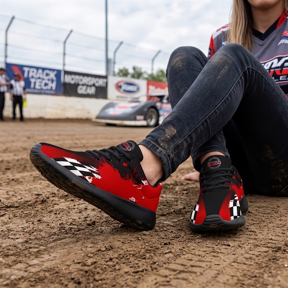 A woman sitting on dirt at a racetrack wearing red and black checkered motorsport sneakers, with a race car and track signage blurred in the background.