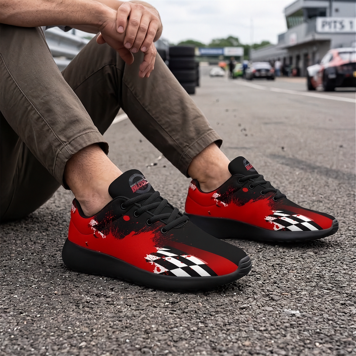 A person sitting on an asphalt racetrack wearing a pair of red and black low-top sneakers with a checkered flag design and red paint-splatter accents. The background shows a blurred racetrack scene with tires and race cars.