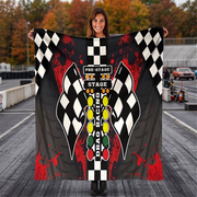 A woman stands on a racetrack holding up a large printed blanket featuring a drag racing theme with checkered flags, red paint splatters, and a starting light design.