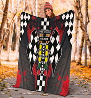 A woman wearing a red beanie holds up a large racing-themed blanket or throw in an outdoor autumn setting with trees and fallen leaves. The blanket features a black and white checkered flag design, red paint splatter graphics, and a central vertical graphic with the words 'PRE-STAGE STAGE' and 'RACING'.