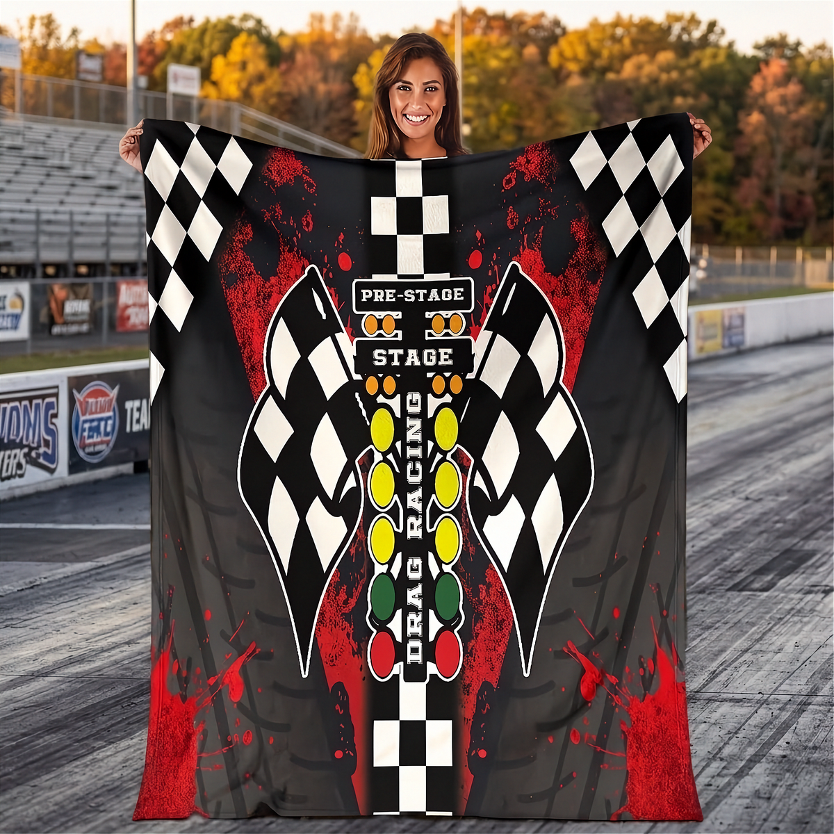 A woman stands on a drag strip holding up a large printed blanket or tapestry featuring a drag racing theme with checkered flags, red paint splatters, and a vertical starting light design.