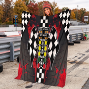 A woman stands on a racetrack holding up a large printed blanket or throw featuring a racing theme with black-and-white checkered flags, red splatter graphics, and a central vertical design with the words 'Pre-Stage Stage Racing' and 'Pga Racing.'.