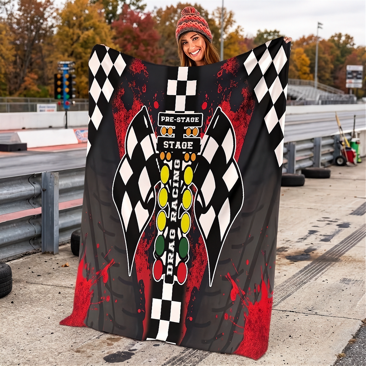 A woman stands on a racetrack holding up a large printed blanket or throw featuring a racing theme with black-and-white checkered flags, red splatter graphics, and a central vertical design with the words 'Pre-Stage Stage Racing' and 'Pga Racing.'.