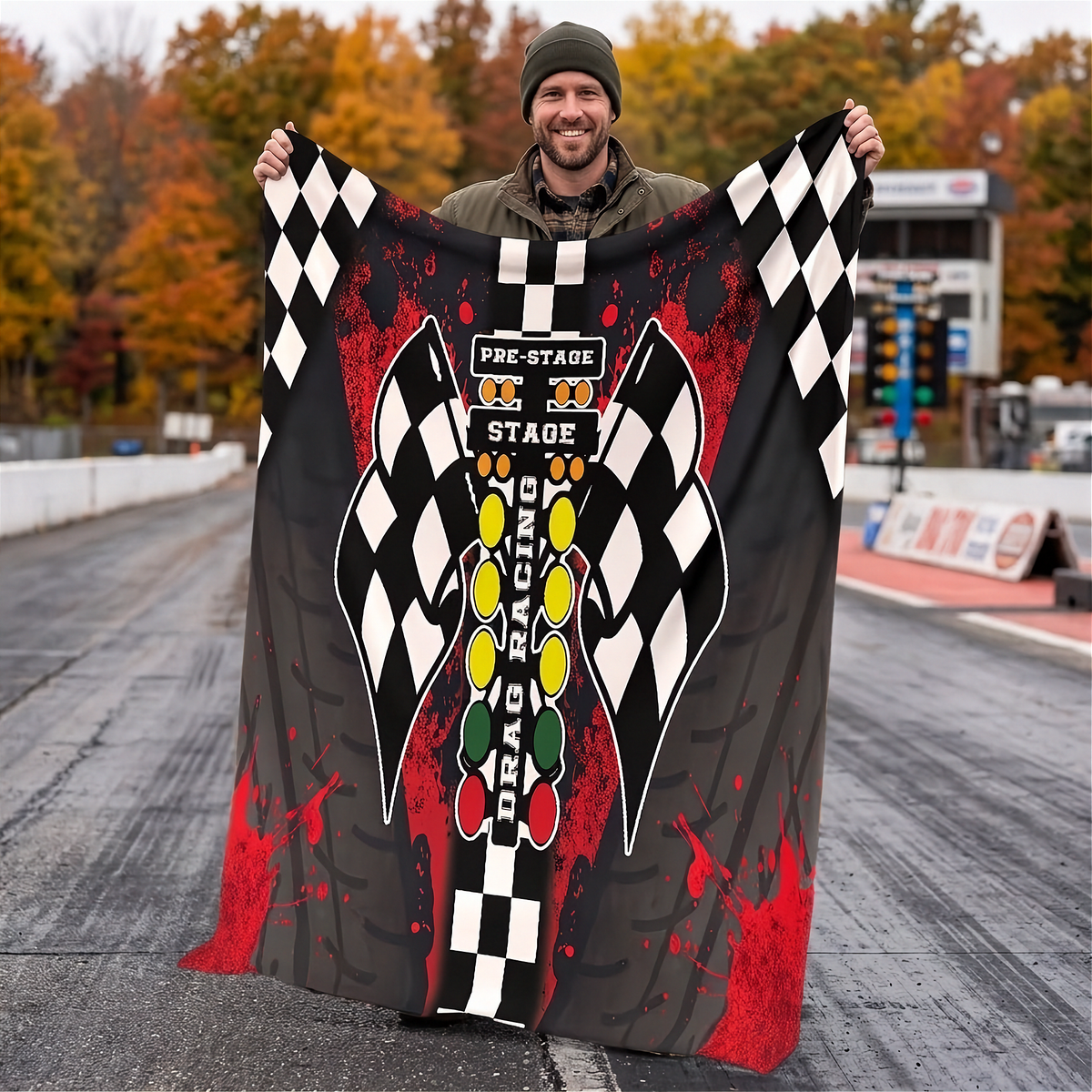 A man standing on a racetrack holds up a large black and gray racing-themed blanket or throw featuring checkered flags, red paint splatter, and a vertical 'Pre-Stage Stage Racing' graphic.