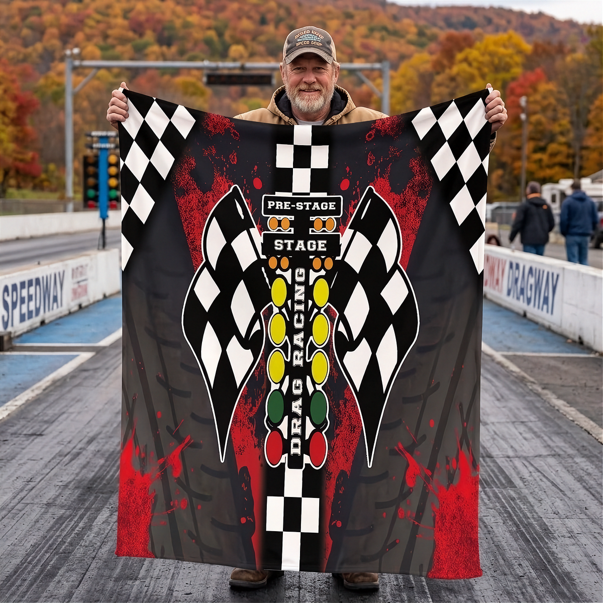 A man stands on a drag strip holding up a large printed blanket or towel featuring a drag racing theme with black-and-white checkered flags, red splatter graphics, and a central vertical graphic with the words 'Pre-Stage 30 Stage Drag Racing.'.