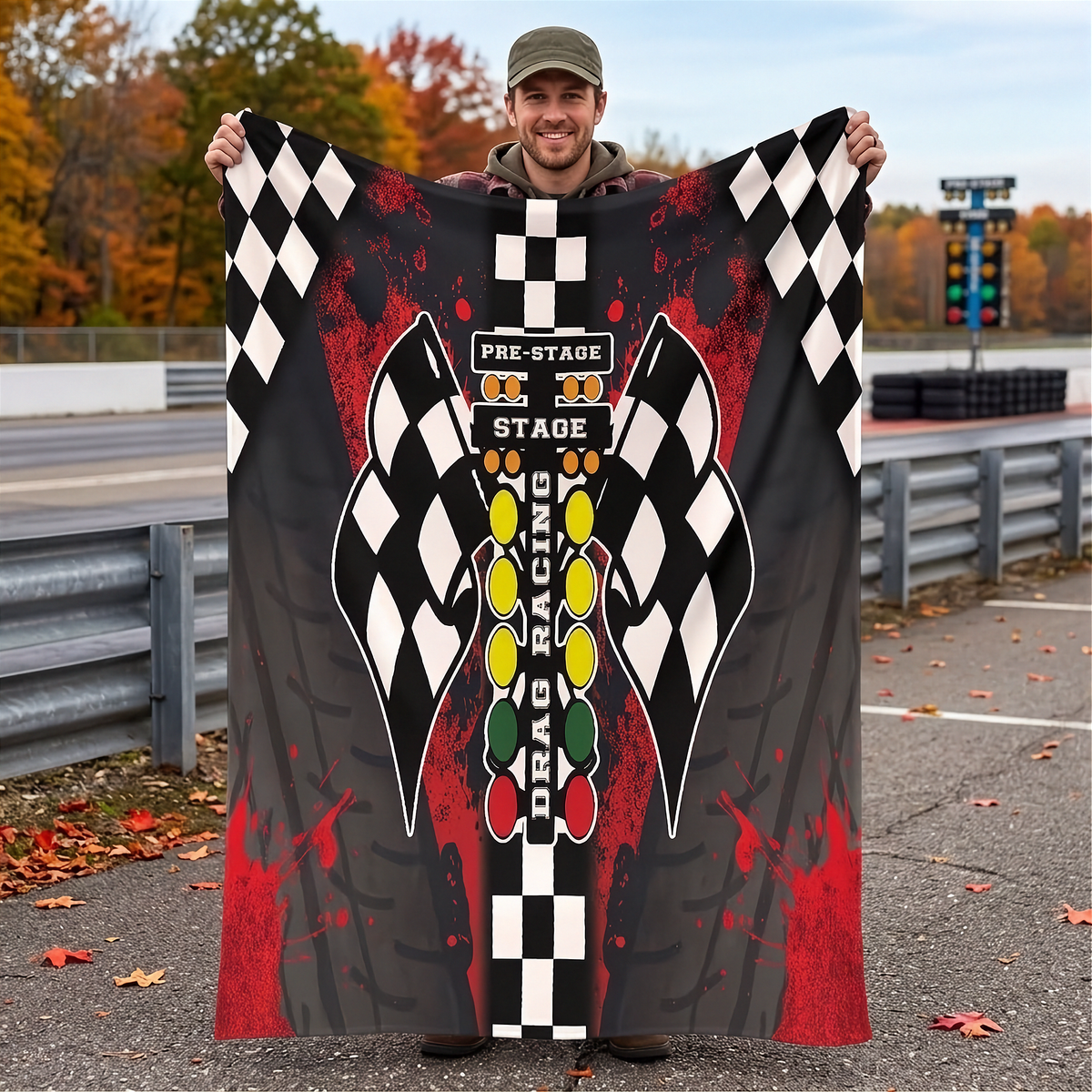 A man wearing a green cap holds up a large printed blanket or poster at an outdoor racetrack. The design features a black and white checkered border, a large wing graphic, and a vertical column of racing lights with the words 'PRE-STAGE', 'STAGE', and 'DRAG RACING'.