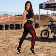 A woman stands on a dirt racetrack wearing black leggings with red 'MOTOCROSS WIFE' text and a heart graphic on the hip, along with a black sports bra. A motocross bike and event tent are visible in the background.